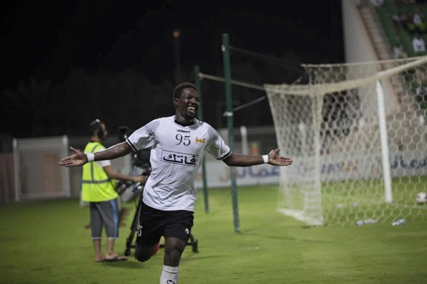 Ein Mann in einem weißen Hemd und schwarzen Shorts rennt über ein Fußballfeld und breitet die Arme aus, umgeben von Zuschauern, mit einem Tor und einer sitzenden Menge im Hintergrund.
