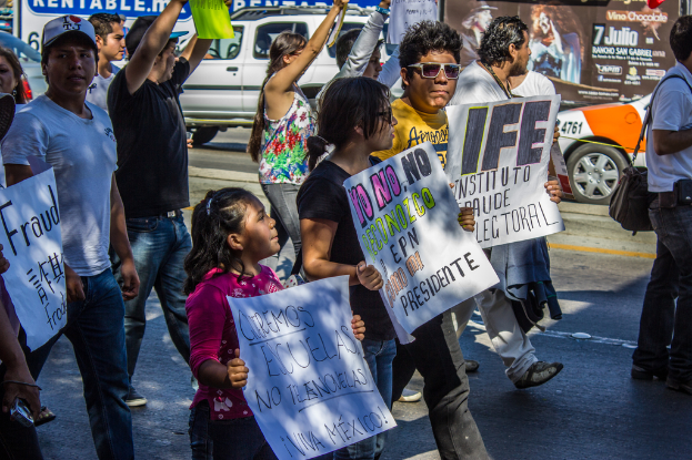 Eine Gruppe von Menschen und Kindern, die Informationsschilder halten, mit einer Straße, Fahrzeugen und Geschäften im Hintergrund.