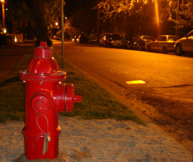 Feuerhydrant am Straßenrand mit Autos und Bäumen im Hintergrund.