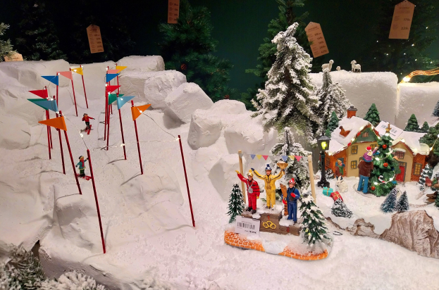 Eine festliche Weihnachtsdorf-Szene mit einem schneebedeckten Berg im Hintergrund, mit Tannen, Häusern, Menschen, Fahnen, Masten, Lichtern, Tieren und anderen Gegenständen, die alle von Schnee bedeckt sind, unter einem dunklen, gemütlichen Ambiente.