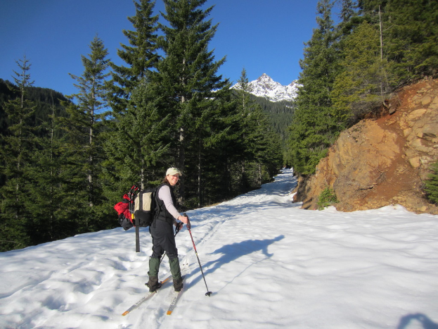 Eine Frau beim Skifahren auf Schnee, mit einem Rucksack und einer Mütze, vor einem Hintergrund aus Bäumen und Hügeln.