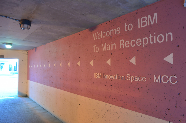 Eine Wand mit dem Text "Willkommen bei IBM", beleuchtet von Deckenlampen, mit einer Tür im Hintergrund, die "IBM Innovationsraum MCC" markiert.
