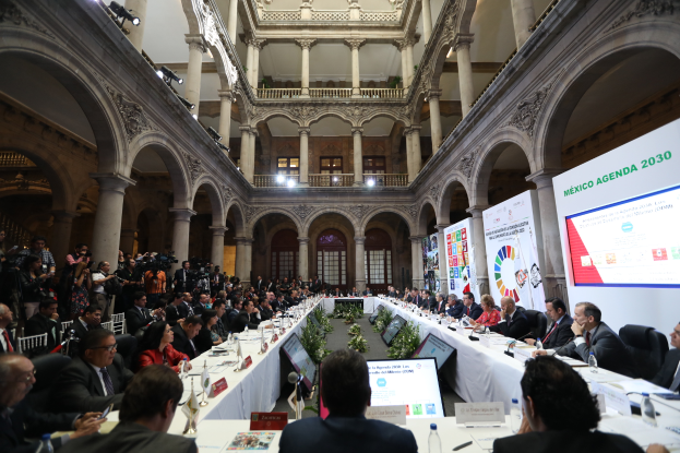 Eine große Gruppe von Menschen sitzt an einem langen Tisch in einem Gebäude, mit verschiedenen Gegenständen auf dem Tisch und Bannern auf der rechten Seite, was auf eine Versammlung für die Mexico-Agenda 2030 hindeutet.