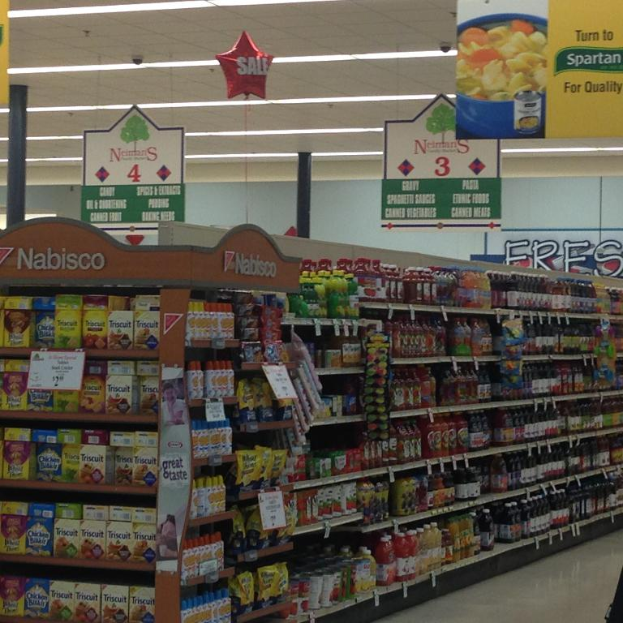 Ein Supermarkt-Innenraum mit Regalen voller Lebensmittel, Deckenlampen und Schildern.