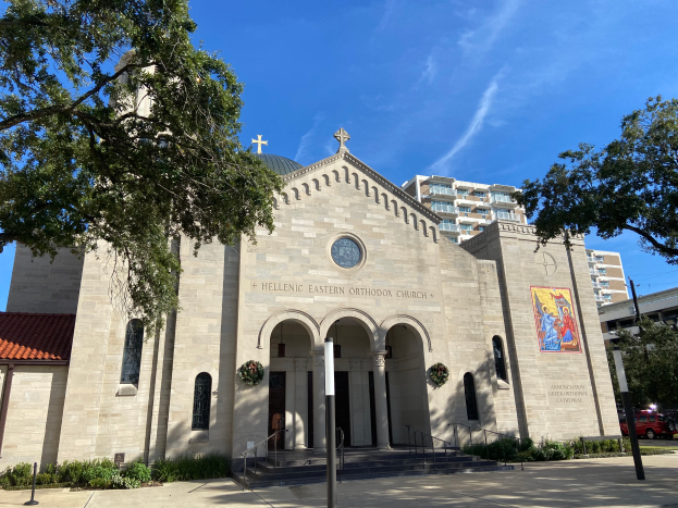 Die griechisch-orthodoxe Kirche in San Antonio, Texas, ist ein großer Bau mit Fenstern, Säulen, Bogengängen, Treppen mit Geländern und einer Wandmalerei, umgeben von Pflanzen, Bäumen und Fahrzeugen auf der Straße, unter einem bewölktem Himmel.