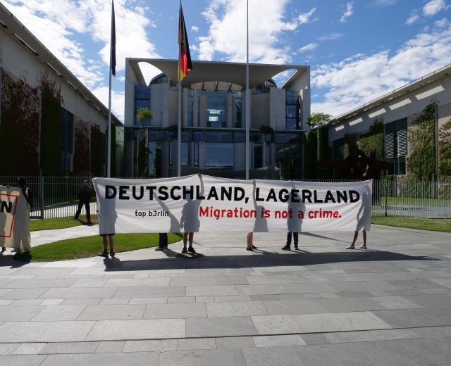 Menschen mit einem Transparent mit der Aufschrift "Deutschland, Lagerland Migration ist kein Verbrechen" vor einem Gebäude mit Fenstern, umgeben von Gras, einem Metallzaun, Pflanzen, Bäumen und Fahnenstangen unter einem bewölkten Himmel.