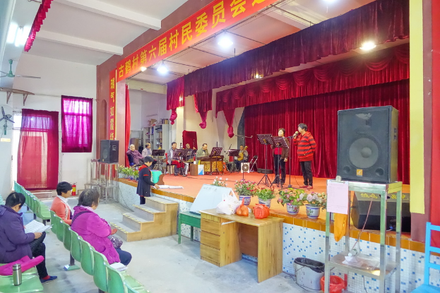 Eine Bühne mit zwei singenden Personen, einer Band im Hintergrund, roten Vorhängen, einem Licht, einem Lautsprecher auf der rechten Seite und einem Publikum auf Stühlen sitzend.