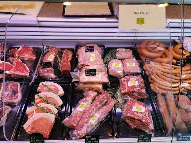 Eine Vitrine voller verschiedener Fleisch- und Wurstwaren in Schalen, jeweils mit Preisschildern, in einer Metzgerei mit einer Tafel im Hintergrund.