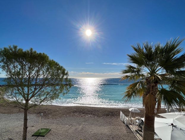 Ein Strand mit Palmen, Sonnenschirmen, üppiger Vegetation, einem strahlend blauen Himmel und einer scheinenden Sonne im Hintergrund, das eine Ferienwohnung in der französischen Riviera zeigt.
