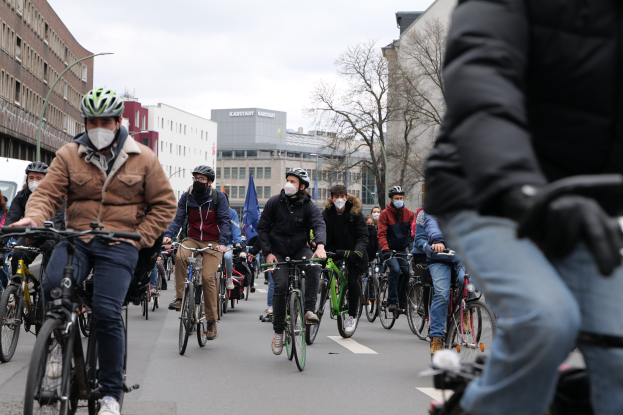 Eine Gruppe von Menschen in Helmen und Handschuhen fährt mit Fahrrädern eine von Bäumen gesäumte Straße in Berlin, Deutschland, entlang, mit Gebäuden und einem geparkten Fahrzeug im Hintergrund.
