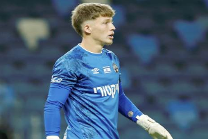 Ein junger Mann in einem blauen Torwart-Outfit, einschließlich eines blauen T-Shirts und weißer Handschuhe, steht auf einem Fußballfeld während eines Spiels, mit einem unscharfen Hintergrund.