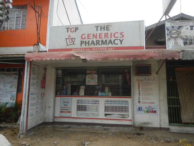 Apothekenfront mit einem "Generika Apotheke"-Schild, umgeben von Gebäuden, Strommasten, Kabeln, Werbetafeln, Grünpflanzen, Fahrzeugen und einem klaren Himmel.