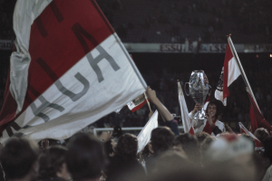 Gruppe von Menschen mit Fahnen und einem Pokal vor einem jubelnden Publikum in einem Stadion.