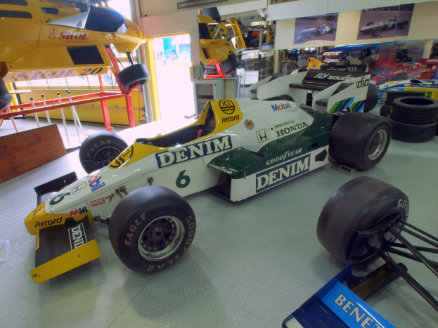 Eine Garage mit verschiedenen Rennwagen, Reifen, einem hölzernen Objekt, einem Geländer, einer Glastür, gerahmten Fotos an einer Wand und Deckenbeleuchtung.