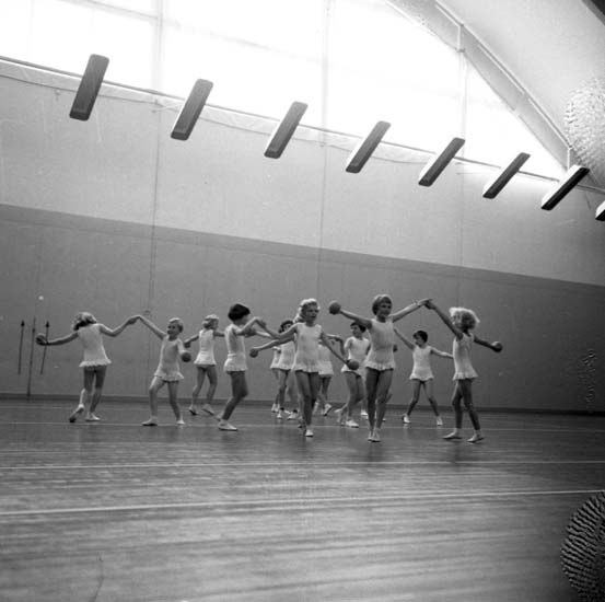 Schwarz-weißes Bild von jungen Mädchen, die in einem Studio tanzen und sich an den Händen halten, mit einem Ball auf der rechten Seite und Lampen an der Decke.