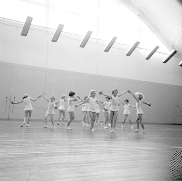 Schwarz-weißes Bild von jungen Mädchen, die in einem Studio tanzen und sich an den Händen halten, mit einem Ball auf der rechten Seite und Lampen an der Decke.