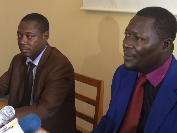 Zwei Männer in Blazern sitzen an einem Tisch mit Mikrofonen, Papieren und anderen Gegenständen und führen ein Gespräch vor einer cremefarbenen Wand mit einem weißen Objekt.