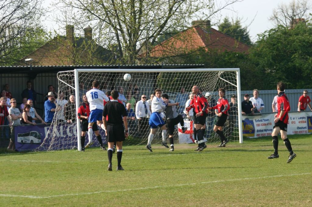 Spieler sind in ein Fußballspiel auf einem Feld mit einem Tornetz involviert, während Zuschauer dahinter stehen, mit Bäumen und Häusern im Hintergrund.