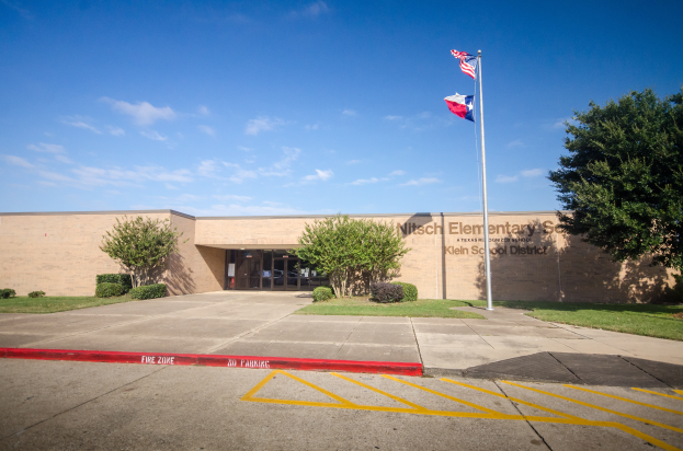 Nilsch-Grundschule mit Glastüren und Text darauf, eine Fahnenstange davor, umgeben von Bäumen, Pflanzen und Gras, mit dem Himmel im Hintergrund.