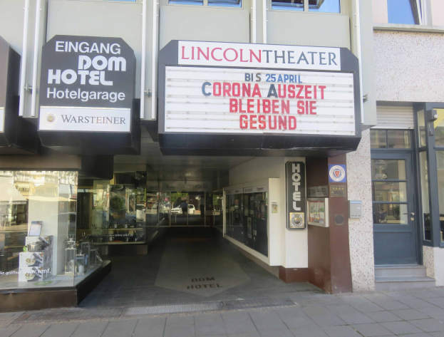 Außenansicht des Lincoln Theaters in Berlin, Deutschland, mit Glasfenstern und -türen sowie einer Schautafel und einem Innenblick, der eine belebte Stadtlandschaft mit verschiedenen Gegenständen zeigt.