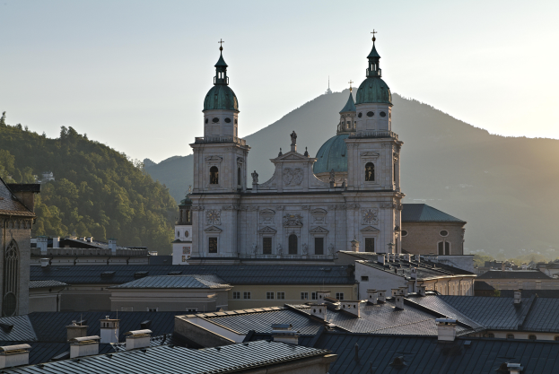 Ein Panoramablick ├╝ber Salzburg, ├ľsterreich, von einem H├╝gel aus, der Geb├Ąude, B├Ąume, eine pr├Ąchtige Kathedrale und ferne Berge unter einem blauen und wei├čen Himmel zeigt.