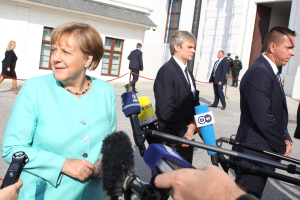 Bundeskanzlerin Angela Merkel spricht vor dem Kanzleramt in Berlin mit Reportern, umgeben von Menschen, einige halten Mikrofone, mit Gebäuden im Hintergrund.