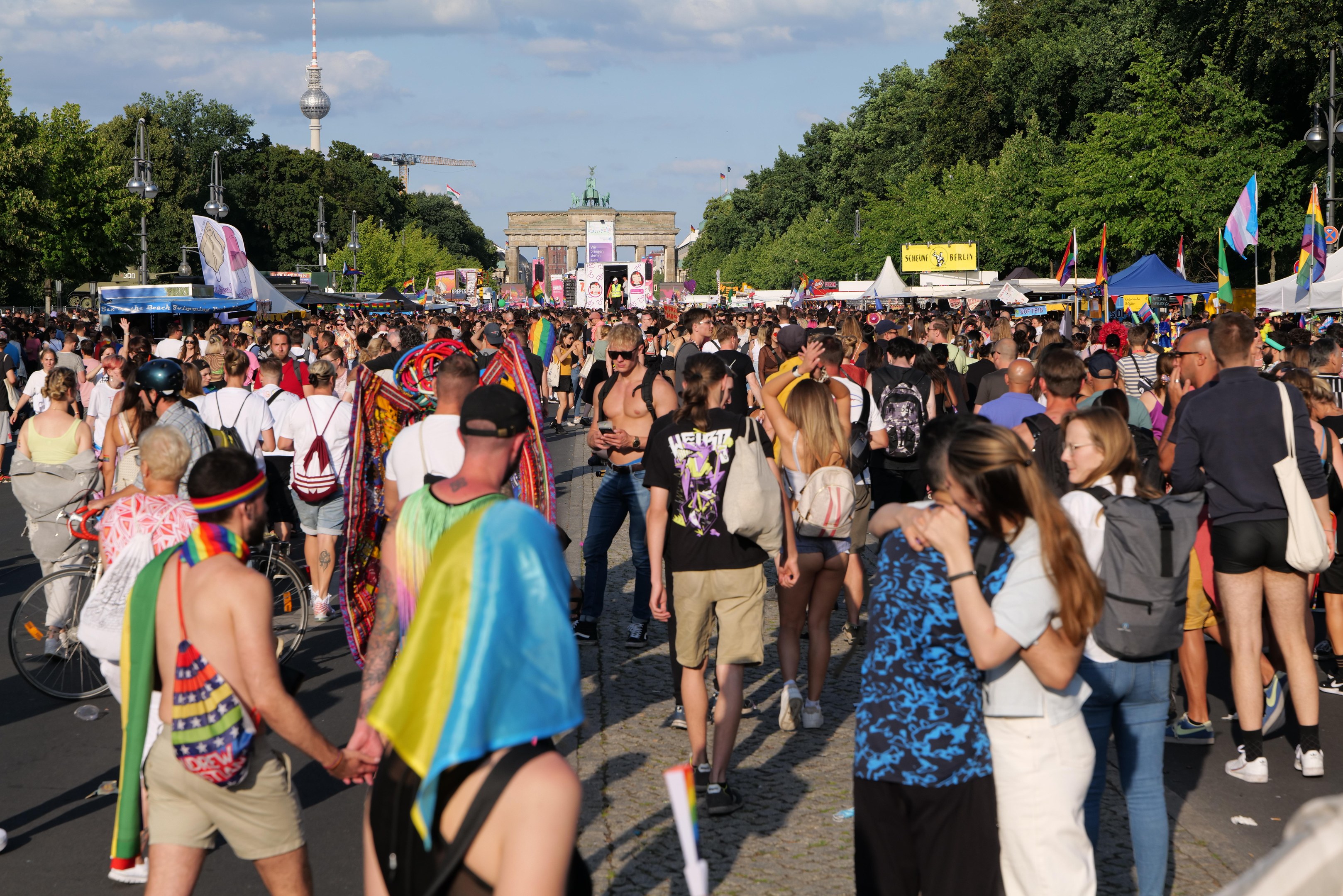 Eine große Menschenmenge marschiert durch eine Straße in Berlin, Deutschland, einige tragen Mützen und tragen Taschen oder Fahnen, mit Zelten, Bannern, Laternenmasten, Bäumen, Gebäuden und einem Turm im Hintergrund unter einem bewölkten Himmel.