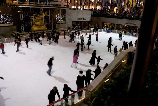 Eine Gruppe von Menschen, die auf einer zugefrorenen Oberfläche Eislaufen, umgeben von Gebäuden.