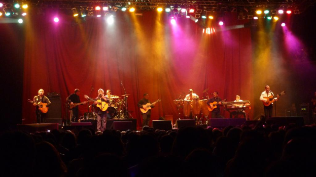 Eine Theateraufführung mit ein paar Leuten, die Gitarren spielen, einem Vorhang im Hintergrund und einer kleinen Gruppe von Menschen am unteren Bildrand.