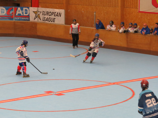 Gruppe von Menschen, die drinnen auf einem Eisstadion Hockey spielen, tragen Helme und Schlittschuhe und halten je einen Hockey-Schläger, mit Bannern und Zuschauern im Hintergrund.