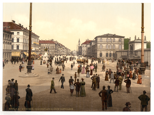 Ein altes Foto einer belebten Münchner Stadtstraße aus den frühen 1900er Jahren, das Fußgänger, einige mit Schirmen, einen Pferdewagen im Vordergrund, Gebäude, Bäume und Pfähle im Hintergrund zeigt, mit Text am unteren Rand des Bildes.