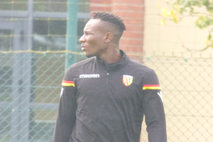 Ein Mann in einer schwarzen Jacke steht auf einem Fussballfeld, umgeben von einem Zaun und einem Baum auf der rechten Seite, mit einem entschlossenen Gesichtsausdruck, scheinbar während eines Trainings.