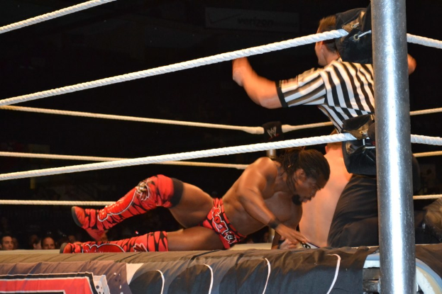 Zwei Männer kämpfen in einem Ring, einer trägt ein rotes Hemd und Schuhe, der andere Shorts, mit einem Schiedsrichter rechts und Zuschauern im Hintergrund unter einem dunklen Himmel.