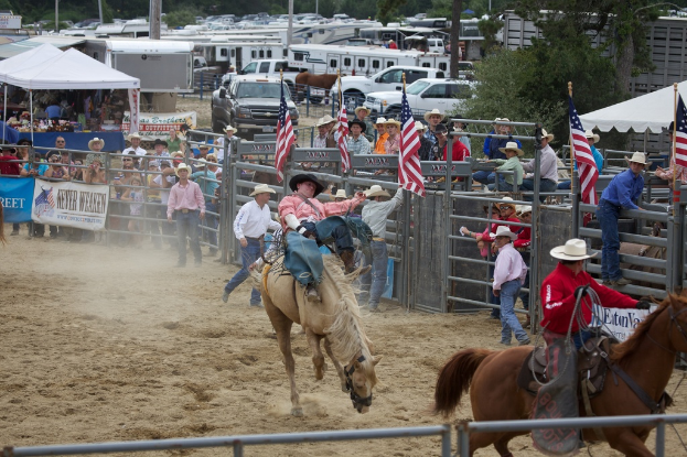 Zwei Reiter auf Pferden bei einem Wettbewerb namens Charreada, umgeben von Gruppen von Menschen, Bannern und Fahnen, Zeltplanen, geparkten Fahrzeugen und Bäumen im Hintergrund.