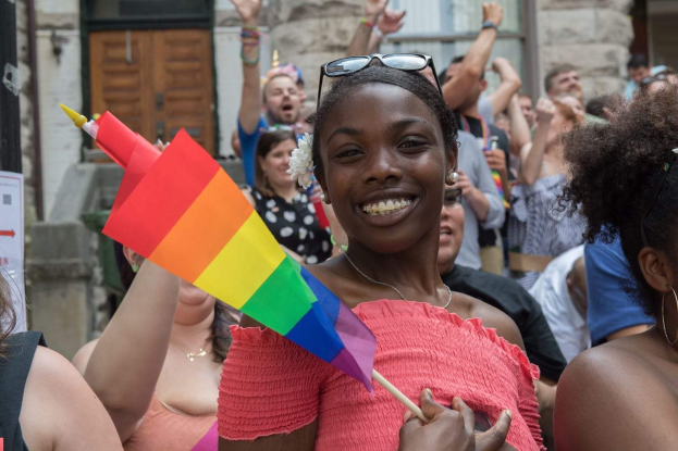 Eine Gruppe von Menschen vor einem Gebäude stehend, mit einer Frau in der Mitte, die eine Regenbogenflagge hält; das Gebäude hat Fenster und eine Tür und ein Schild mit Text ist sichtbar.