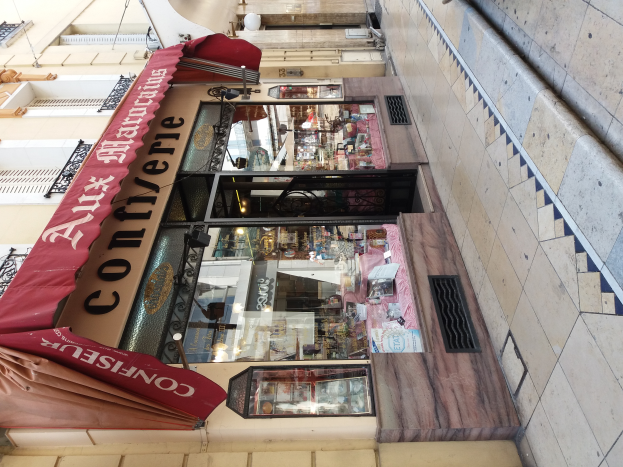 Ein Schaufenster mit einer roten Markise, umgeben von Gebäuden mit Fenstern, Geländern und Lichtern, das verschiedene Gegenstände im Inneren zeigt, die darauf hindeuten, dass es ein Confiserie-Shop ist.