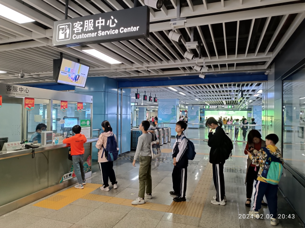 Gruppe von Menschen, die an einem Flughafen-Check-in-Schalter mit Glaswänden auf der rechten Seite, Monitoren und einer Service-Informationstafel darüber stehen.