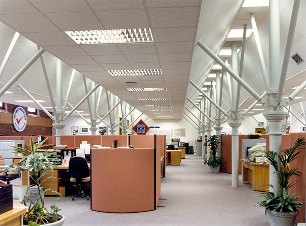 Ein leerer Büroraum mit mehreren Tischen und Stühlen, die ordentlich angeordnet sind, Topfpflanzen für Grün, Säulen, eine Wanduhr und Deckenleuchten.