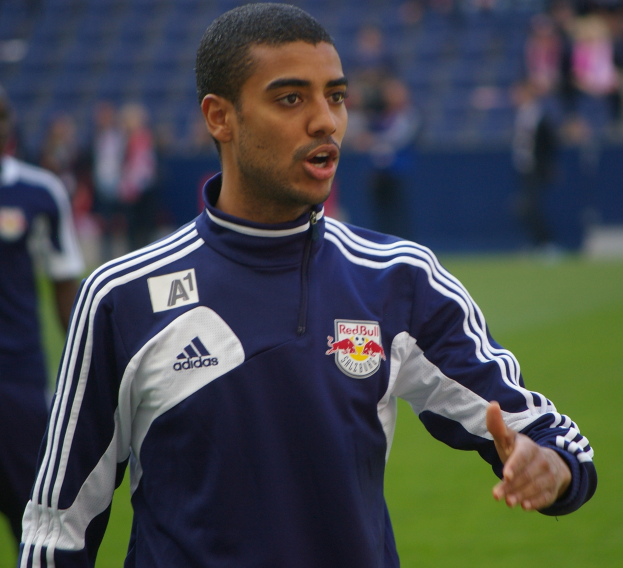 Ein Mann in einer blauen Jacke steht auf einem Fussballfeld, umgeben von einer Gruppe von Menschen, mit einem entschlossenen Gesichtsausdruck.