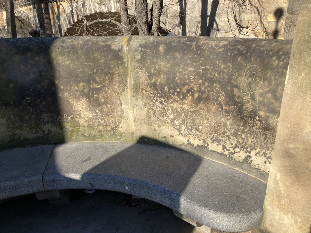 Eine alte Steinbank in einem Park, umgeben von einer Mauer, Bäumen und einem Gebäude im Hintergrund.