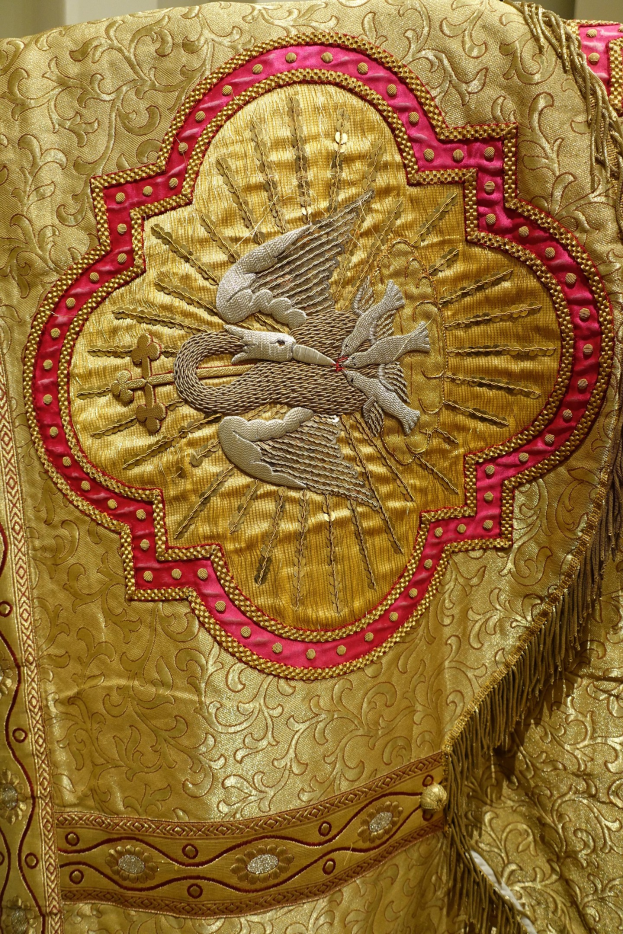 Nahaufnahme eines Priestergewandes mit goldenen und roten Stickereien und detaillierten Mustern vor einem neutralen Hintergrund.