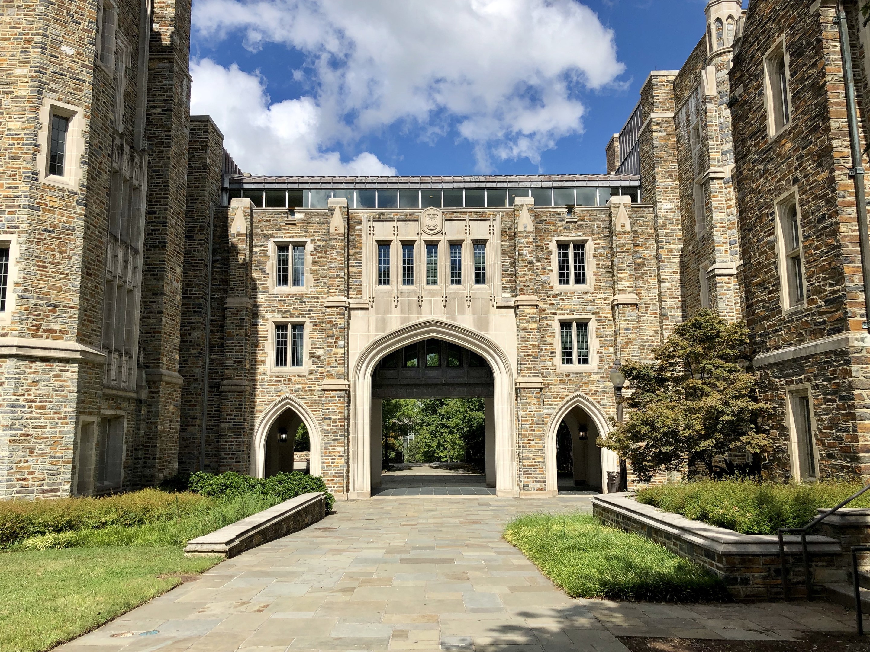 Großes steinernes Universitätsgebäude mit gewölbtem Eingang, umgeben von grünem Gras, Pflanzen, Bäumen und einem bewölkten Himmel.