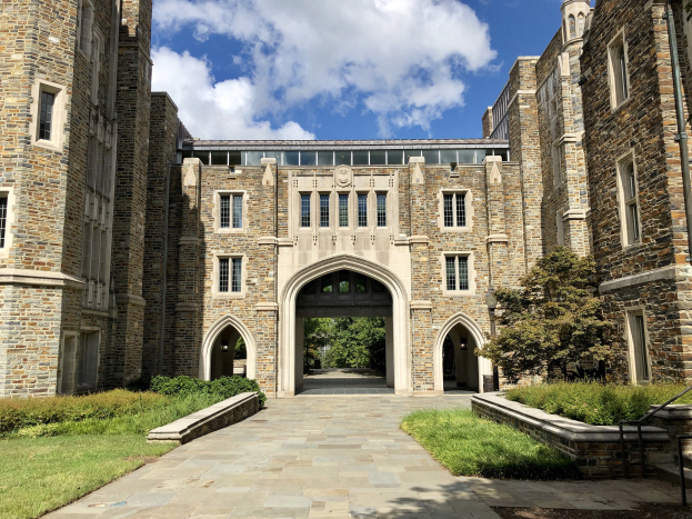 Großes steinernes Universitätsgebäude mit gewölbtem Eingang, umgeben von grünem Gras, Pflanzen, Bäumen und einem bewölkten Himmel.