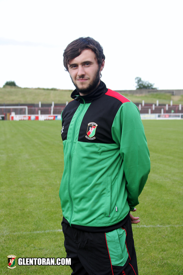 Ein Mann in einer grünen Jacke steht auf Gras mit einem Zaun und einem Torpfosten im Hintergrund, Bäume und Himmel darüber.