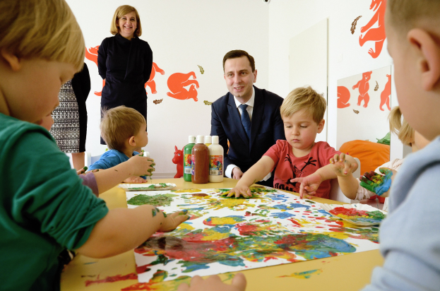 Kinder bei einer Malaktivität an einem Tisch mit farbigen Händen, Papieren und Flaschen, beaufsichtigt von einer lächelnden Frau, mit Kunstwerken an der Wand.