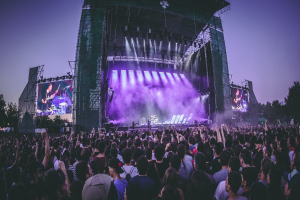 Eine große Menge Menschen bei einem Musikfestival schaut auf einer hell erleuchteten Bühne mit Instrumenten und Bildschirmen, die vor einem Gebäude, Bäumen und einem klaren blauen Himmel steht.