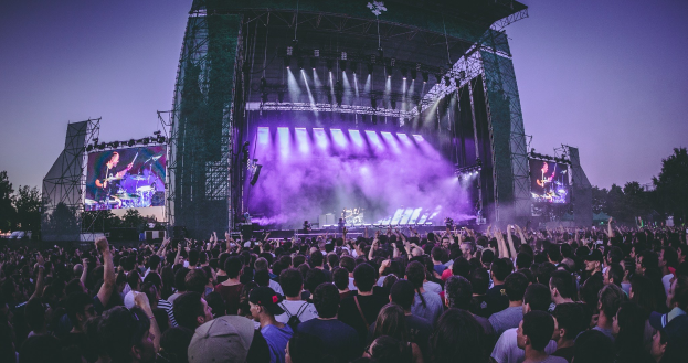 Eine große Menge Menschen bei einem Musikfestival schaut auf einer hell erleuchteten Bühne mit Instrumenten und Bildschirmen, die vor einem Gebäude, Bäumen und einem klaren blauen Himmel steht.