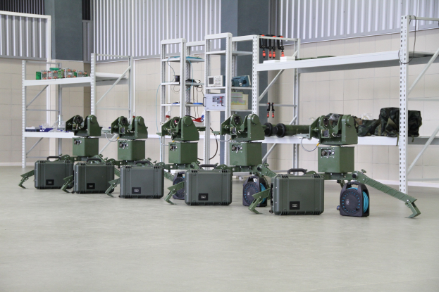 Eine Gruppe von Militärrobotern auf einem Boden mit Koffern und Gegenständen auf Gestellen, vor einer Wand mit einem Geländer im Hintergrund.