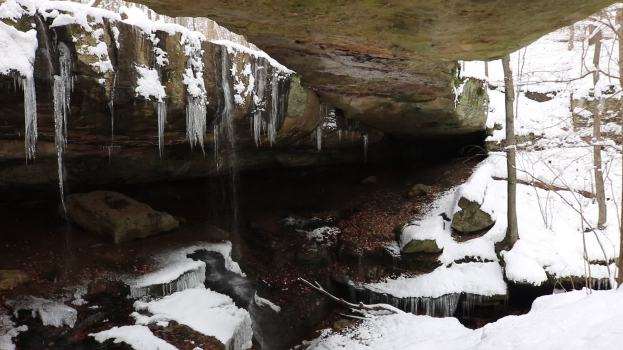 Ein kleiner Wasserfall ergießt sich über einen schneebedeckten, felsigen Abhang in einem bewaldeten Gebiet, mit Eiszapfen an den Felsen.