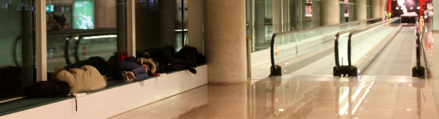Menschen auf Bänken in einem Flughafen mit zwei geraden Rolltreppen im Hintergrund.